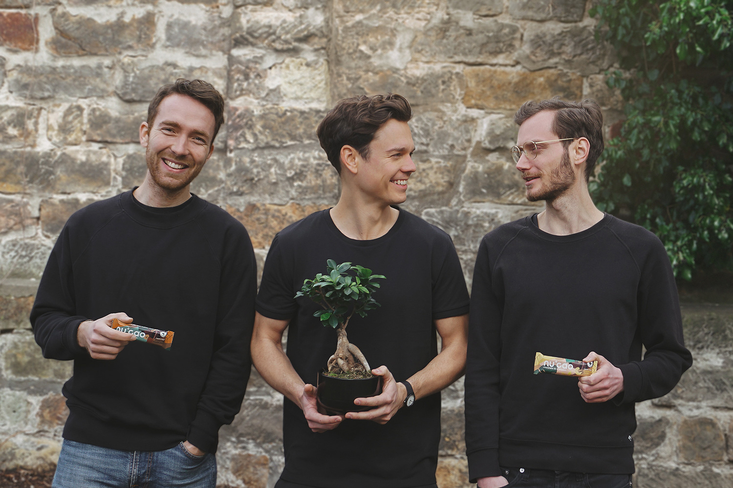 drie jonge mannen voor een muur met mueslireep in de hand en een plant die op een boom lijkt