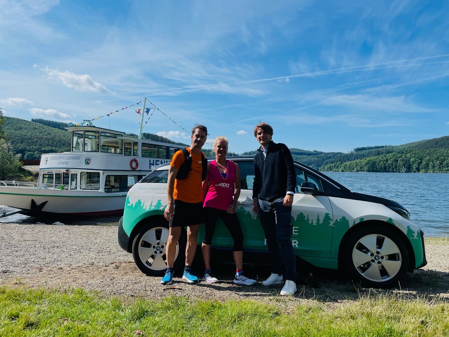 Drei Personen vor einem E-Auto stehen vor einem See. 